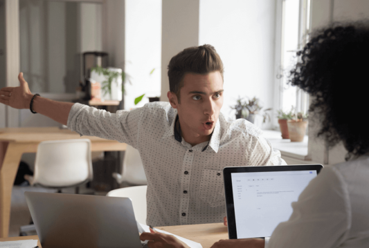 Two employees sitting across from one another at a desk with a laptop between them looking like they are disagreeing about something