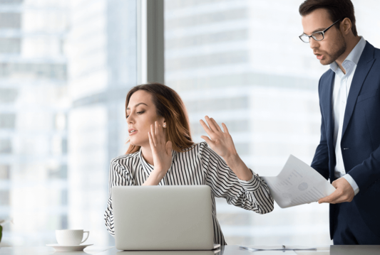 Woman in front of laptop with her hands up trying to block man over her shoulder trying to show her a document