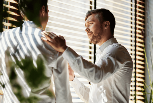 Two employees by the window talking. One employee has one hand on the other employees shoulder