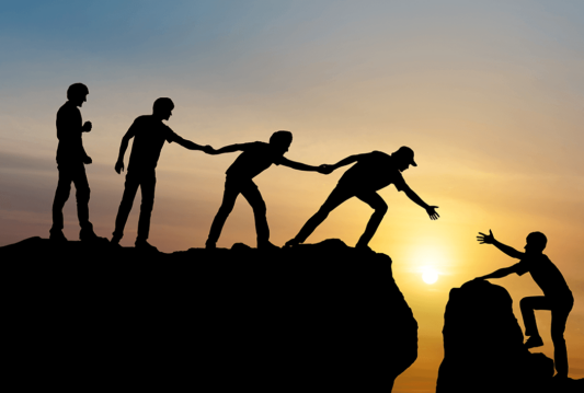 4 people on a cliff at sunset holding hands in a line tying to help lift up a fifth person on the other side of the cliff