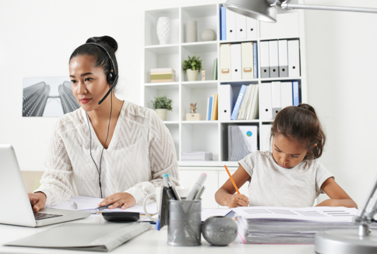 A mother working on her laptop beside her daughter doing her home work