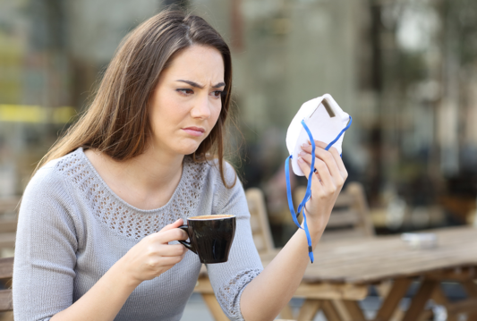 A woman sitting down having a coffee and staring at a facial mask in her other hand