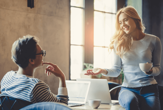 an employee sitting at a desk and another employee sitting on the desk with a coffee flirting with one another