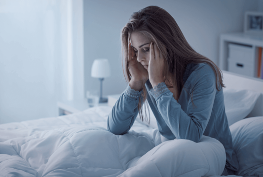 A woman sitting on her bed in her pajamas rubbing her temples